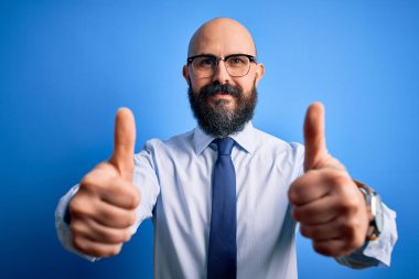 Sakallı, zarif kravat takan, mavi arka planda gözlük takan, el hareketini onaylayan, gülümseyen ve başarı için mutlu bir iş adamı. Kazanan jest.