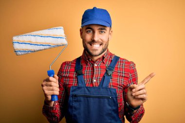 Genç yakışıklı ressam duvarları yalıtılmış sarı arka planda silindir kullanarak boyuyor. Çok mutlu bir şekilde el ve parmak uçlarını işaret ediyor.