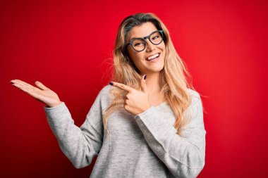 İzole edilmiş kırmızı arka planda süveter ve gözlük takan genç güzel sarışın kadın hayrete düşmüş ve kameraya gülümserken eliyle ve parmağıyla işaret ediyordu..