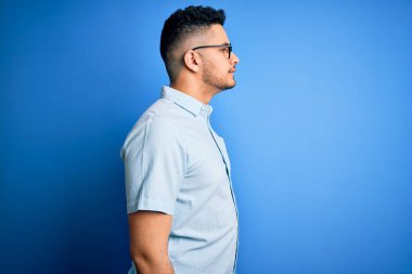 Genç ve yakışıklı bir adam, günlük tişört ve gözlük takıyor, mavi arka planda, yan tarafa bakıyor, rahat profil pozu veriyor, kendine güvenen bir gülümsemesi var..