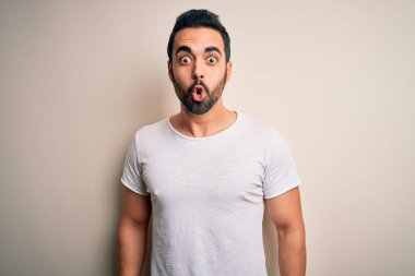 Sakallı, sıradan tişörtlü genç bir adam beyaz arka plan üzerinde korku dolu ve şaşkınlık dolu bir ifade, korku ve heyecanlı bir yüz ifadesiyle duruyor..