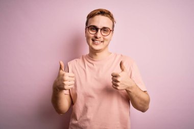 Genç, kızıl saçlı, günlük tişört giyen, pembe arka plan başarı tabelasının üzerinde duran, elle olumlu jest yapan, gülümseyen ve mutlu genç bir adam. Neşeli ifade ve kazanan jest.