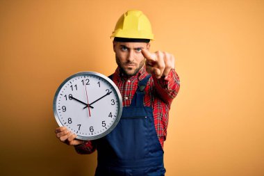 Güvenlik kaskı takan genç adam sarı arka plan üzerinde büyük bir saat tutuyor. Kamerayı işaret ediyor ve sen, el işareti, ön taraftan pozitif ve kendinden emin bir hareket.