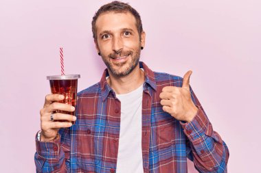 Genç yakışıklı adam bir bardak kola içiyor mutlu ve pozitif gülümsüyor, başparmağı mükemmel ve onay işareti yapıyor. 