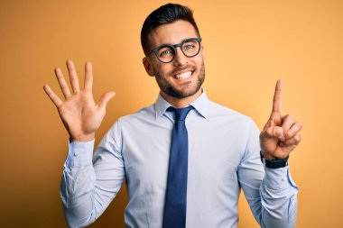 Sarı arka plan üzerinde duran kravat ve gözlük takan genç yakışıklı işadamı altı numaralı parmaklarıyla gülümseyerek kendinden emin ve mutlu görünüyor..