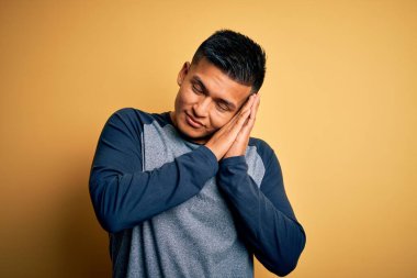 Genç, yakışıklı, latin tişörtlü bir adam sarı arka planda dikiliyor. Yorgun uyuyor, hayal kuruyor, elleriyle poz veriyor ve gözleri kapalı gülümsüyor..