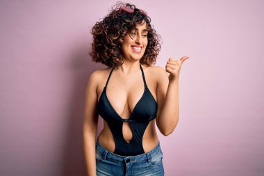 Genç, güzel bir Arap kadın, mayo ve pembe arka planda güneş gözlüğü takmış, gülümseyen, gülümseyen ve baş parmağıyla yüzünü gösteren bir şekilde tatilde..