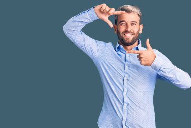 Zarif bir gömlek giyen yakışıklı sarışın adam gülümseyerek elleri ve parmakları gülen yüzlü bir çerçeve çiziyor. yaratıcılık ve fotoğrafçılık kavramı. 