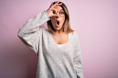 Genç, güzel, esmer bir kadın pembe arka planda günlük süveter ve gözlük takıyor. İnançsız ifade.