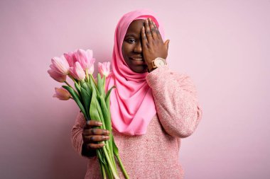 Genç, Afro-Amerikan, iri yarı, Müslüman bir kadın elinde pembe bir buket lale ile bir gözünü kapatıyor, yüzünde kendine güvenen bir gülümseme ve şaşırtıcı bir duygu var..