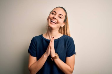 Mavi gözlü genç, güzel, sarışın bir kadın beyaz arka plan üzerine günlük tişörtler giyip ellerini birleştirip af diliyor kendinden emin bir şekilde gülümsüyor..