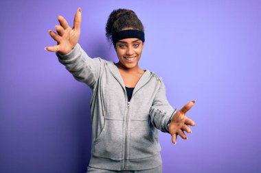 Genç Afro-Amerikan sporcu kadın spor yapıyor mor arka plan üzerine spor giyerek kucaklaşmak için kollarını açıp gülümseyen kameraya bakıyor. Mutluluğu kucaklayan neşeli bir ifade..