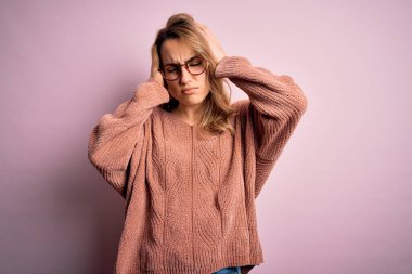 Genç, güzel, sarışın bir kadın pembe arka plan üzerinde gözlük ve süveter takıyor. Ağrı ve migren yüzünden baş ağrısı ve stres altında. Ellerini başının üstüne koy..
