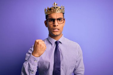 Genç ve yakışıklı Afro-Amerikalı adam mor arka planda kralın altın tacını takıyor kızgın ve kızgın yumruğunu kaldırıyor ve öfkeyle bağırıyor. Öfke ve agresif kavram.