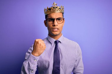 Young handsome african american man wearing golden crown of king over purple background angry and mad raising fist frustrated and furious while shouting with anger. Rage and aggressive concept. clipart