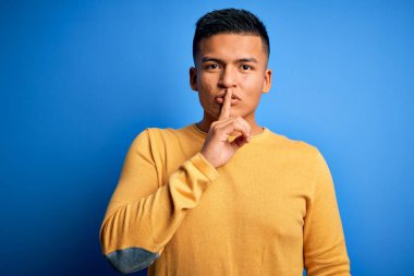 Sarı günlük süveter giyen genç, yakışıklı latin adam izole edilmiş mavi arka planda sessiz olmak istiyor. Sessizlik ve gizli konsept.