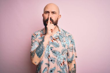 Sakallı, dövmeli, pembe arka planda sıradan bir çiçek gömleği giyen yakışıklı kel adam sessiz olup dudaklarını öpmek istiyor. Sessizlik ve gizli konsept.