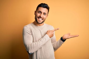 İzole edilmiş sarı arka plan üzerinde duran gündelik süveter giyen genç yakışıklı adam kameraya hayret ediyor ve gülümsüyor..