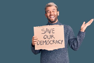 Genç sarışın adam demokrasi kartonu pankartımızı kurtardı zaferi mutlu bir gülümseme ve kazanan bir ifadeyle kutluyor ellerini kaldırarak 