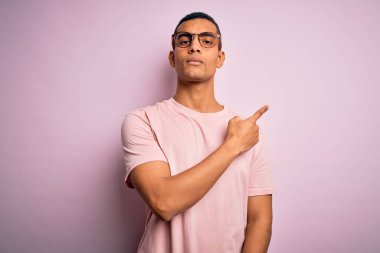 Yakışıklı Afro-Amerikalı adam pembe arka planda günlük tişört ve gözlük takıyor. Elleriyle işaret ediyor. Ciddi ve sakin bir yüz ifadesi sergiliyor.