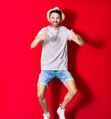 Genç, yakışıklı, günlük kıyafetler giyen ve şapkalı mutlu bir İspanyol. Yüzünde tebessümle zıplıyor. Tamam işareti yapıyor. Kırmızı arka plan üzerinde başparmaklar yukarı doğru..