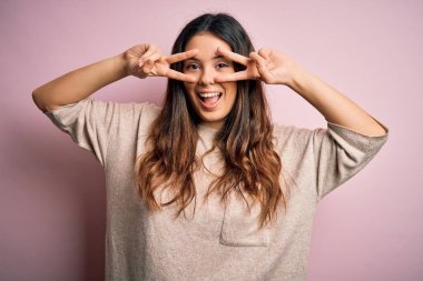 Genç, güzel esmer kadın pembe arka planda dikilip barış sembolü yapıyor. Yüzünde parmaklarıyla gülümsüyor ve zaferini gülümsüyor.
