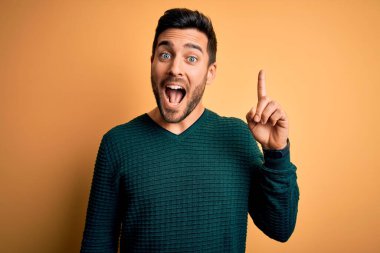 Sarı arka planda dikilmiş, sakallı, gündelik süveter giyen genç ve yakışıklı bir adam başarılı bir fikirle parmağını kaldırıyor. Heyecanlı ve mutlu. Bir numara..