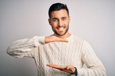 İzole edilmiş beyaz arka plan üzerinde duran, büyük ve büyük boyut işareti gösteren, sıradan bir süveter giyen genç bir adam. Kameraya bakıp gülümsüyor. Ölçüm konsepti.
