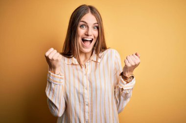 İzole edilmiş sarı arka plan üzerinde sıradan çizgili tişört giyen genç, güzel, kızıl saçlı kadın kollarını kaldırarak ve açık gözlerle başarıyı kutluyor ve şaşırıyor. Kazanan konsept.