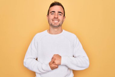 Mavi gözlü genç adam sarı arka plan üzerinde rahat ve neşeli gülümseyerek ellerini çapraz yaparak duran sıradan bir süveter giyiyor. Başarı ve iyimser