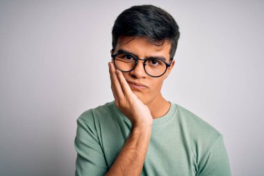 Genç ve yakışıklı bir adam günlük tişört ve gözlük takıyor. İzole edilmiş beyaz arka plan. Kolları çaprazlanmış depresyondan sıkılmış ve yorgun görünüyor..