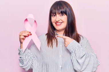 Elinde pembe kanser kurdelesi tutan genç ve cüsseli bir kadın gülümsüyor. 