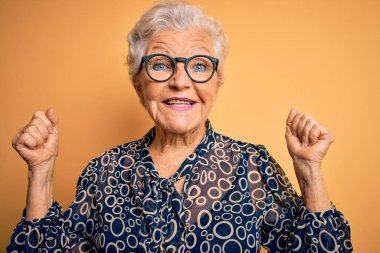 Sarı arkaplan üzerine gündelik gömlek ve gözlük takan son sınıf güzeli gri saçlı kadın kollarını kaldırarak ve açık gözlerle başarıyı kutluyor ve şaşırıyor. Kazanan konsept.