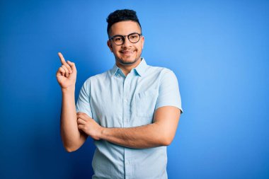 Genç ve yakışıklı bir adam günlük tişört ve gözlük takıyor mavi arka planda, yüzünde kocaman bir gülümseme, eli ve parmağı kameraya bakıyor..