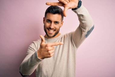 İzole edilmiş pembe arka plan üzerinde duran sıradan bir süveter giyen genç yakışıklı gülümseyerek elleri ve parmakları gülen yüzlü bir çerçeve yapıyor. Yaratıcılık ve fotoğrafçılık kavramı.
