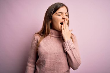 Güzel genç bir kadın pembe izole edilmiş arka plan üzerine balıkçı yaka kazak giyiyor. Elle ağzını kapatmaktan sıkılmış. Huzursuz ve uykulu..