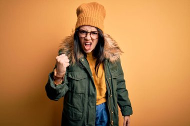 Genç esmer kadın, gözlük takıyor ve sarı şapkalı kışlık bir ceket giyiyor. İzole edilmiş arka plan kızgın ve kızgın. Öfkeyle bağırırken yumruğunu kaldırıyor. Öfke ve agresif kavram.