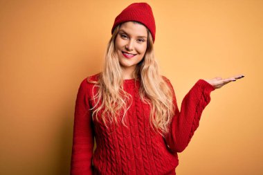 Beyaz arka planda günlük kazak ve yün şapka giyen genç, güzel sarışın kadın mutlu bir şekilde sunum yapıyor ve avuç içi kameraya bakıyor..