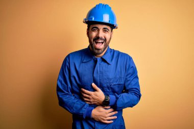 Sakallı tamirci mavi üniformalı ve kasklı sarı arka planda gülümseyen ve kahkahalarla gülen adam..