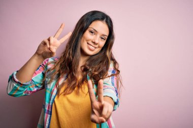 Genç, güzel, esmer, sıradan bir gömlek giyen pembe arka planda durup parmaklarını zafer işareti yaparken gösteren kameraya gülümseyen bir kadın. İki numara..