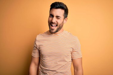 Sakallı, sarı arka planda sıradan çizgili tişört giyen yakışıklı genç adam kameraya seksi bir ifadeyle bakıyor, neşeli ve mutlu bir yüzle..