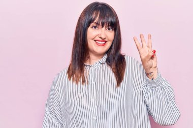 Genç ve cüsseli kadın rahat kıyafetler giyip üç numaralı parmaklarıyla işaret ederken kendinden emin ve mutlu gülümsüyor.. 