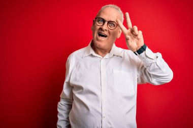 Orta yaşlı, yakışıklı, beyaz gömlekli, kırmızı arka planda gözlüklü, barış sembolü yapan, gülümseyen, zaferi gösteren bir adam.