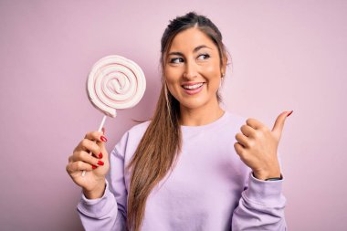 Genç, güzel esmer kadın izole edilmiş pembe arka planda şeker yiyor ve gülümseyen yüzüyle baş parmağıyla işaret ediyor.