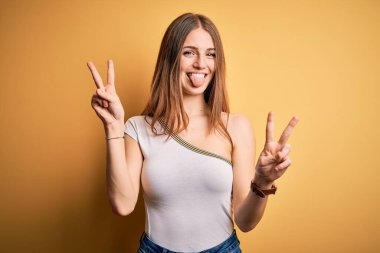 Genç, güzel, kızıl saçlı bir kadın izole edilmiş sarı arka plan üzerine günlük tişört giyip iki elini de zafer işareti yaparken gösteren bir dille gülümsüyor. İki numara..