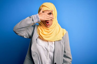 Orta yaşlı esmer iş kadını, geleneksel Müslüman tesettüre bürünmüş mavi arka planda, yüzü ve gözleri kapalı, utangaç bir ifadeyle parmaklarının arasından bakıyor..
