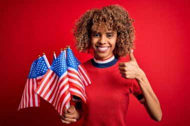 Genç Afro-Amerikan vatansever kıvırcık kadın kırmızı arka planda bayrakları tutuyor. Gülümsüyor ve gülümsüyor. İşaret veriyor.