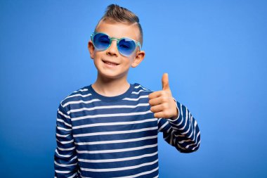 Mavi gözlü, mavi arka planda güneş gözlüğü takan, mutlu bir el hareketi yapan küçük beyaz bir çocuk. Kameraya bakıp başarıyı gösteren ifade onaylanıyor.