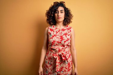 Genç, kıvırcık Arap bir kadın tatilde. Çiçekli bir elbise ve güneş gözlüğü takıyor. Yüzünde ciddi bir ifadeyle rahatlıyor. Basit ve doğal bir şekilde kameraya bakıyorsun..