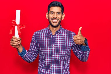 Diploması olan genç Latin bir adam mutlu ve pozitif gülümsüyor, başparmağı mükemmel ve onay işareti yapıyor. 